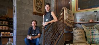 Daniel and Monica Garland pose with cups of coffee on a staircase near sacks of coffee beans. By David Zickl