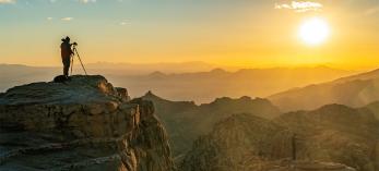 Photographer with tripod standing on rock cliff looking toward sunset.