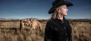 Deborah Copenhaver Fellows looks off into the distance standing in tall grass with her horse a few steps away. By Joel Grimes