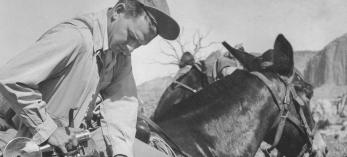 BW photo of Ray Manley lifting a 4x5 camera out of a bag while mounted on a mule in the desert. | COURTESY OF CAROLYN MANLEY ROBINSON