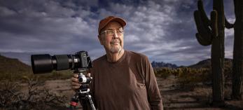 Jack Dykinga poses under a cloudy sky in the desert with a long-lensed camera mounted on a tripod. | JOEL GRIMES 