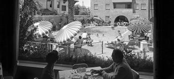 A black and white image shows two people, a woman and a man, in shadow at a dining table within the interior of the Westward Ho hotel in downtown Phoenix. The pair looks out onto a busy swimming pool, surrounded by striped sunbrellas. 