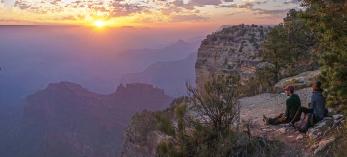 Two people watch the sun set from the edge of the Grand Canyon.