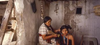 One indigenous woman sits on a crate, fixing another indigenous woman's hair. 