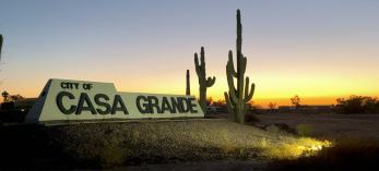 A sign welcoming visitors to Casa Grande is framed by saguaros and lit by spotlights.