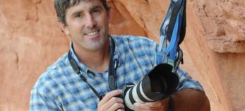 A color photograph shows a smiling white man in a blue plaid shirt. He is anchored to a rock wall with climbing gear and holding a camera with a large lens. 