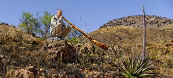 Photograph of man crouching on a rock in the desert blowing into a didgeridoo is by Steven Meckler