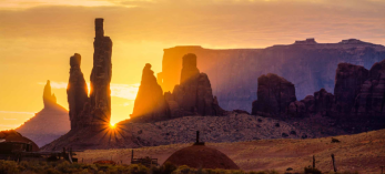 The sun peeks between the iconic spires of Monument Valley Navajo Tribal Park. By David Muench