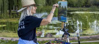 Photograph of a woman painting from an easel outside by a pond is by John Burcham